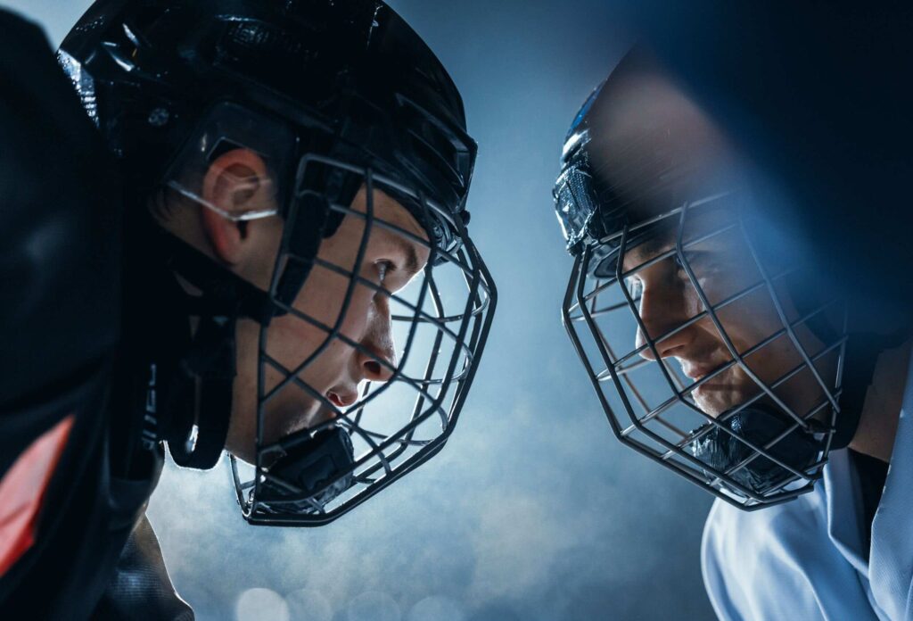 2 hockey players from different teams wearing hockey masks face each other and look at each other during a puck drop