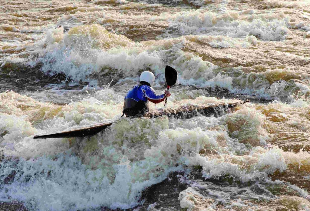 A man in a white helmet and blue suit navigates a strong grade 4 whitewater current in a kayak