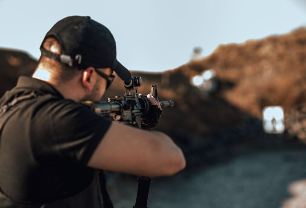 A man in a black polo and black cap aims a rifle through a telescopic sight at a target at a far distance