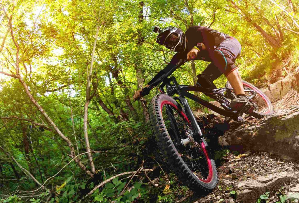 A man on a black bicycle with a red rim, wearing equipment and a helmet with an action camera, goes down the rocks on a forest trail