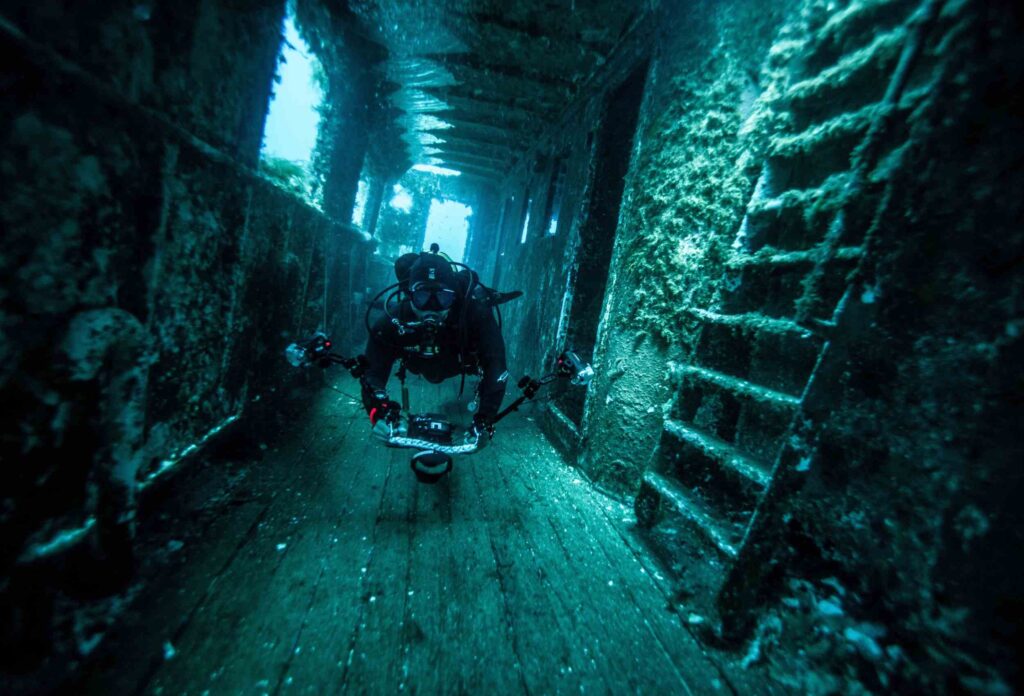 A diver with professional equipment is diving in a sunken ship, swimming along a corridor that is overgrown with algae
