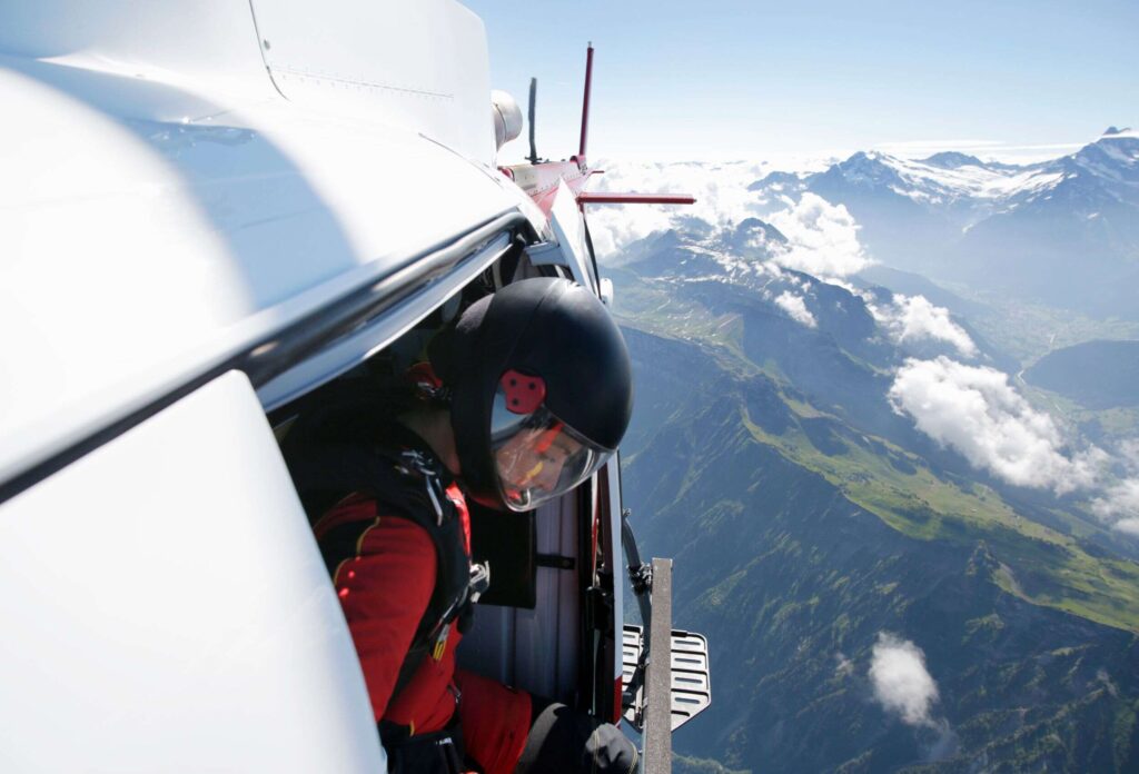 photo of a man standing at the open door of a helicopter hovering in the air preparing for a parachute jump from a helicopter against the background of beautiful snowy mountain ranges