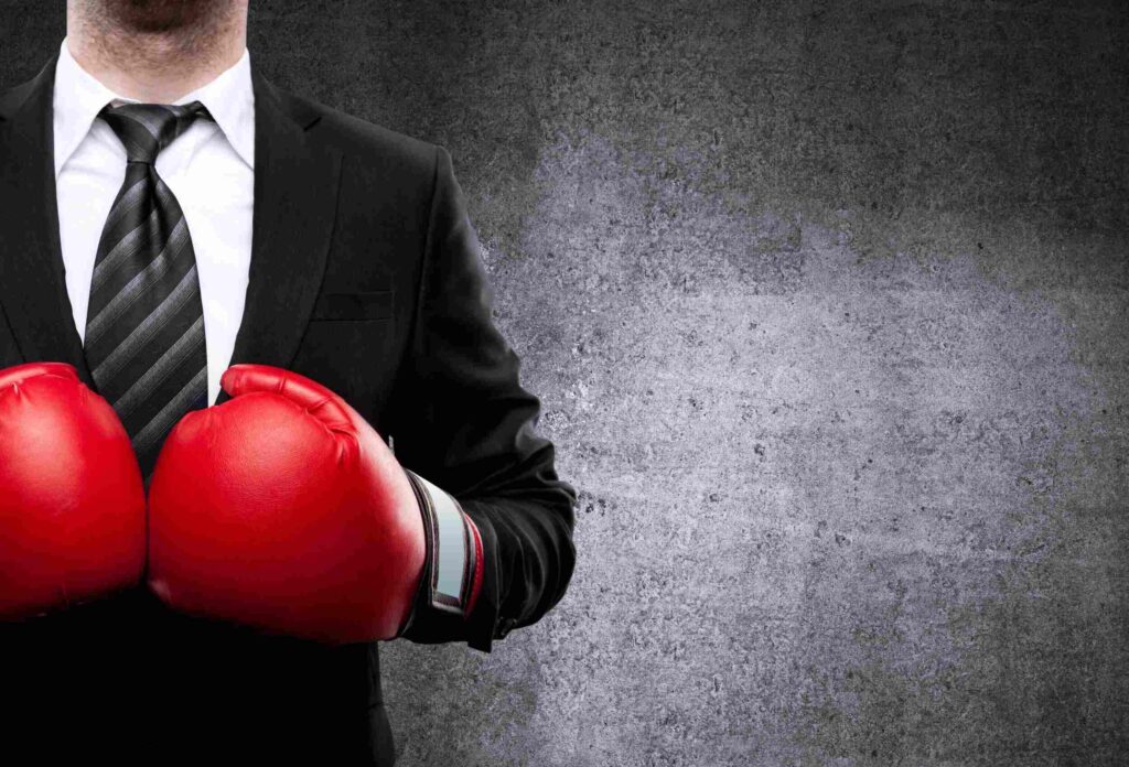 A photo of a man in a black suit, white shirt and tie standing against a gray background, wearing red boxing gloves