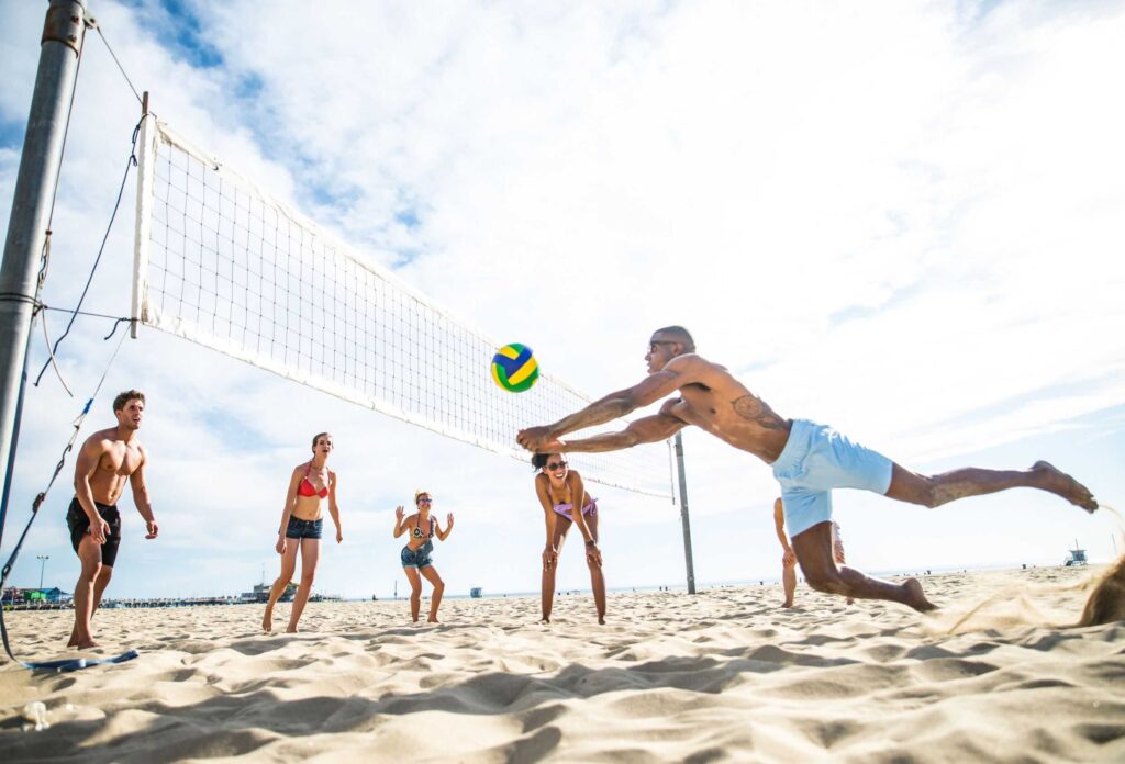 A bald man in white shorts, glasses and tattoos makes a dangerous dive for the ball during a game of beach volleyball