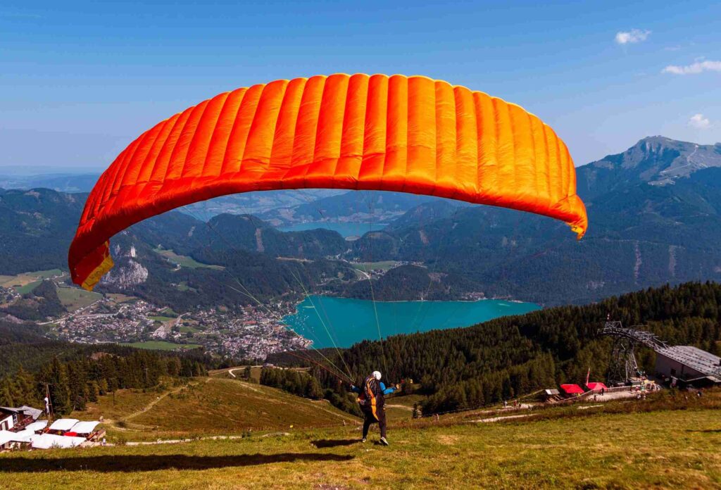 A paraglider accelerates down a mountainside and performs a flight in a bright orange paraglider against a backdrop of a lake town and a pine forest