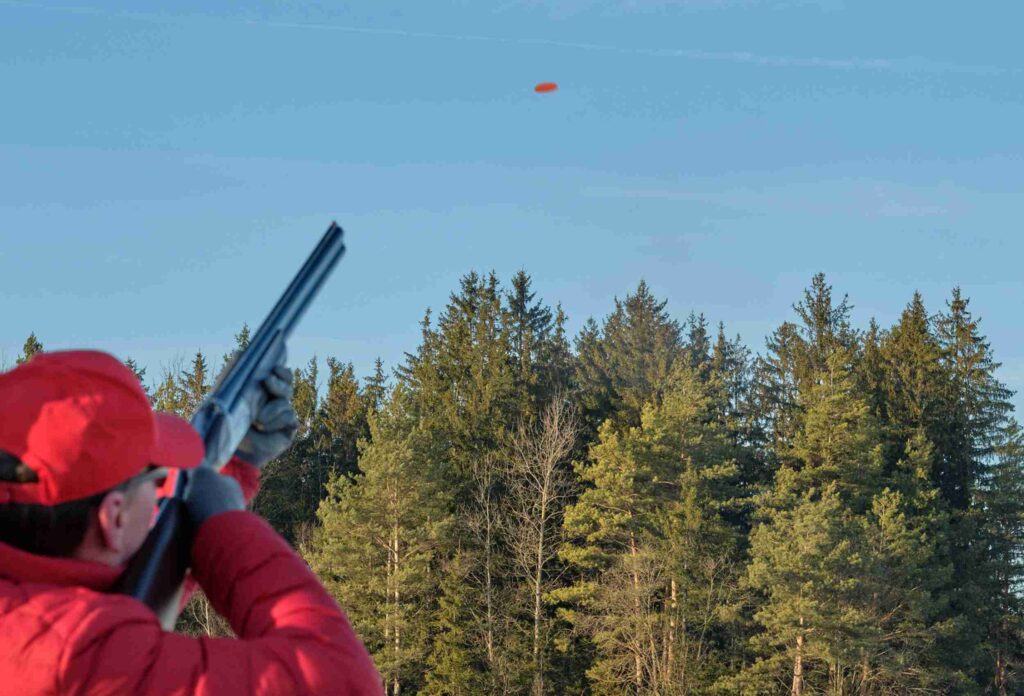 a man in a red jacket and red cap aims a shotgun at a high-flying clay pigeon against a background of green spruce trees