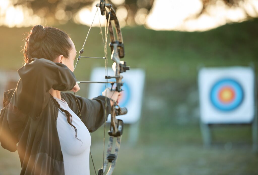 a girl with black hair braided into a braid in a white T-shirt and a black cape pulls the bowstring with an arrow and aims with a crossbow at the target