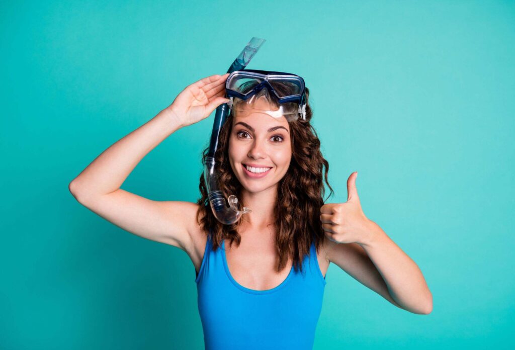 a girl in a blue swimsuit with a snorkel and diving goggles shows an "ok" gesture.