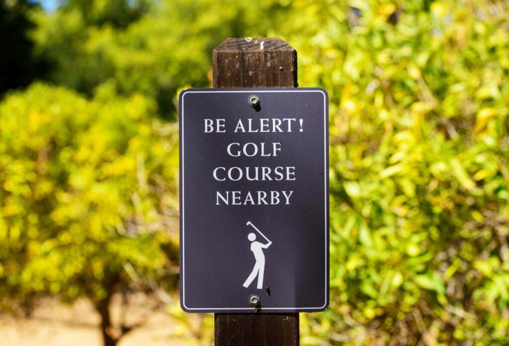There is a black sign in the park with a picture of a man playing golf and a warning to be careful as there is a golf course nearby