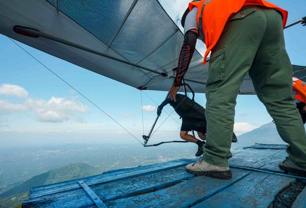 A hang glider accelerates on a wooden bridge on top of a mountain against a backdrop of marvelous views, assisted by an instructor in an orange suit