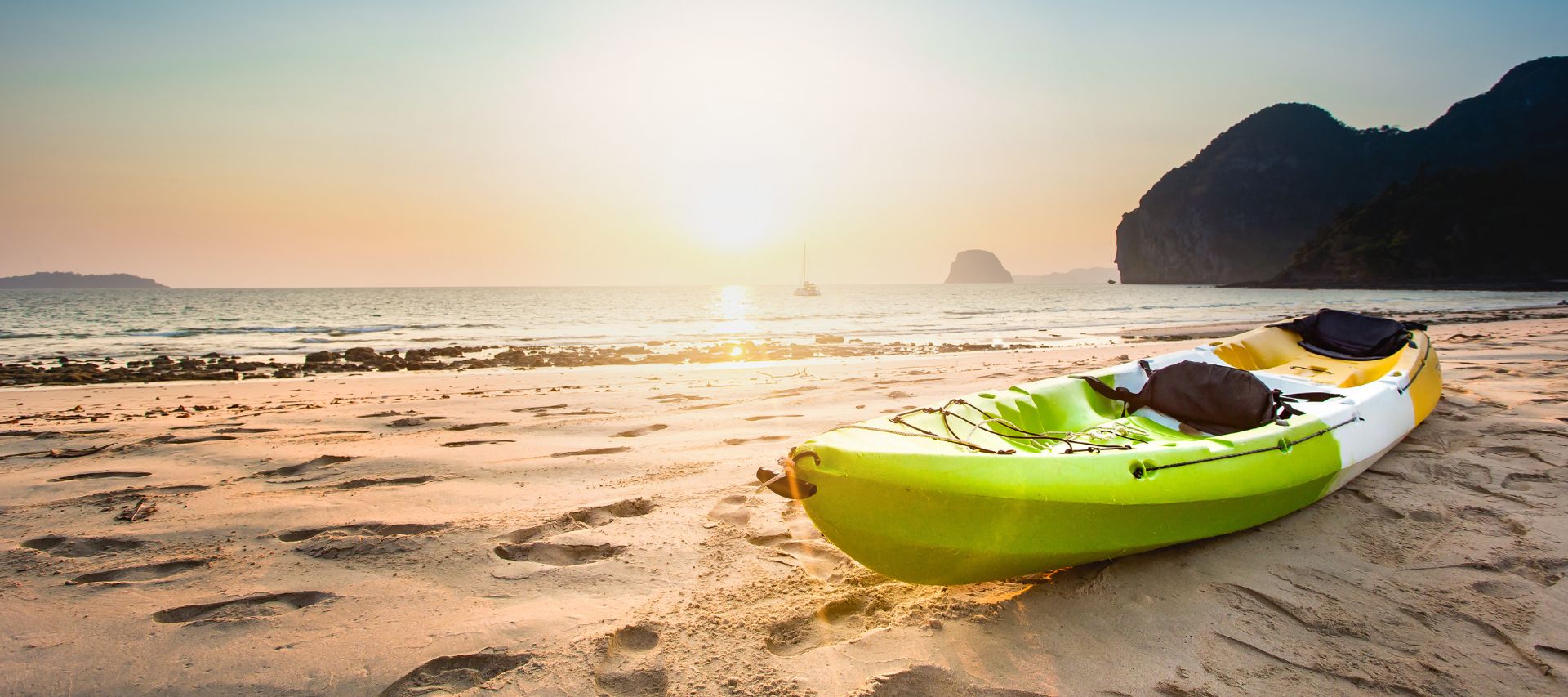 A tri-colored kayak in light green, white and yellow lies on the beach against a beautiful sunset and mountain backdrop