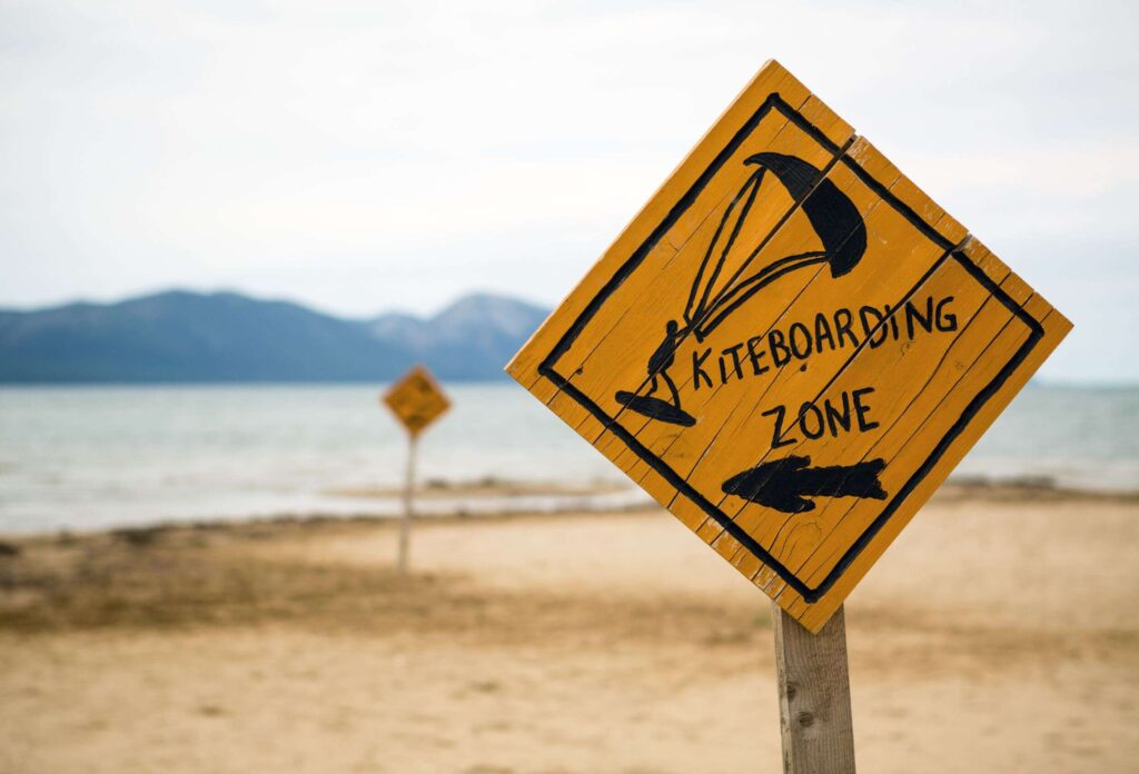 There is a black and orange sign in the sand on the beach that says "kiteboarding zone" and an arrow pointing in the right direction