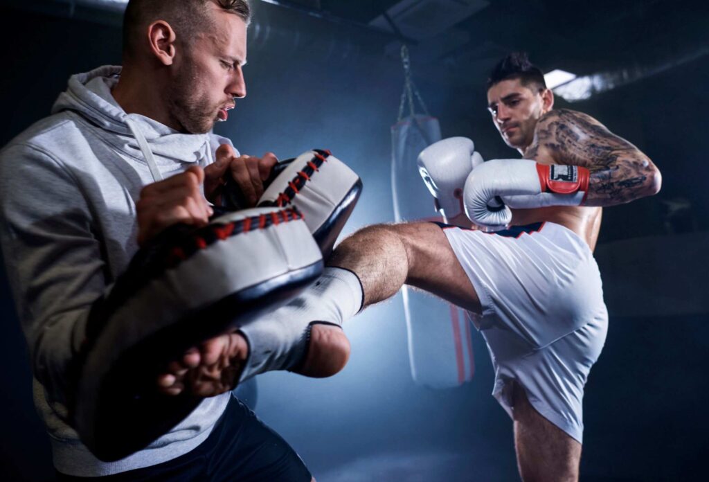 A man with a tattoo on his arm wearing white gloves kicks his leg during a workout with a trainer