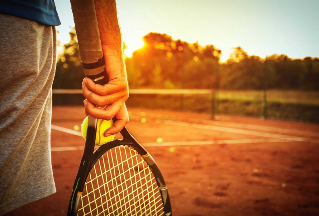 A photograph from the back of a man standing on a tennis court during sunset and holding a racket and ball in his hand