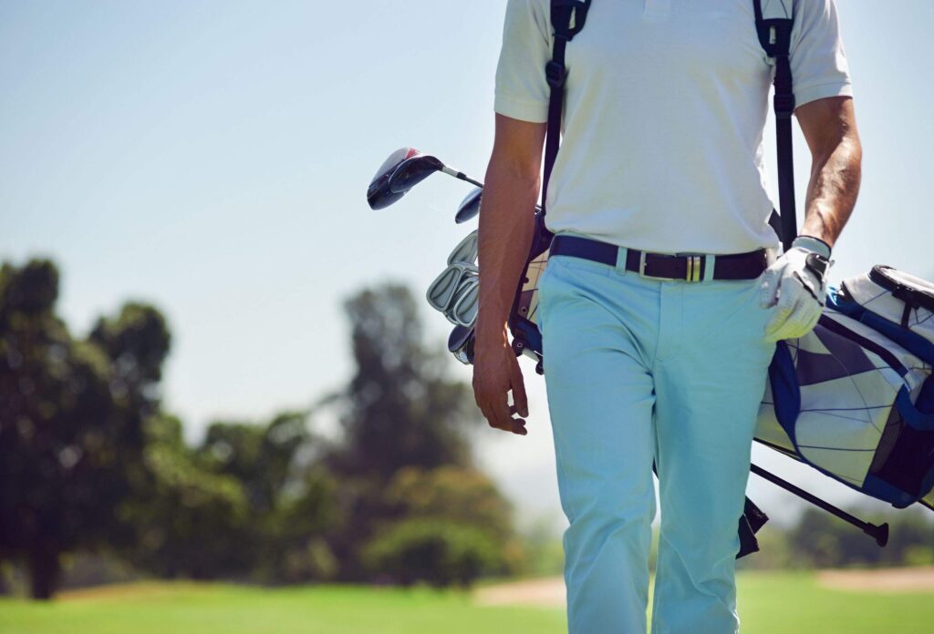 stylish tanned man in blue pants, golf glove and white T-shirt walking along the golf course carrying golf clubs in a golf bag