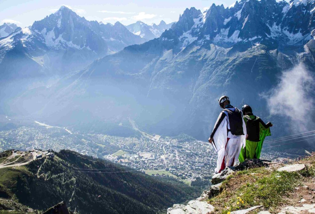 2 men in white and green wingsuits stand on the edge of a cliff and prepare to jump against a backdrop of green and snowy mountains with a city between them