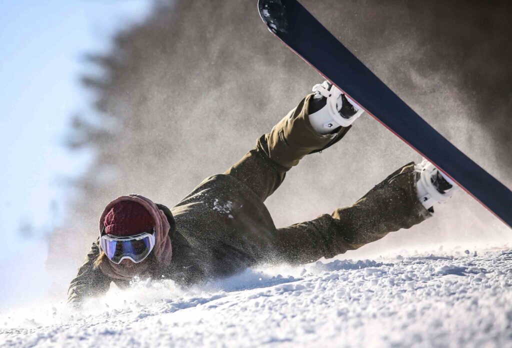 A snowboarder in a red knitted hat and khaki suit dynamically falls on her stomach