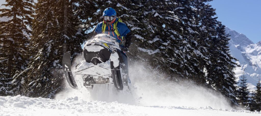 A man in a blue suit with a backpack makes a jump on a snowmobile against the background of trees and snowy mountains