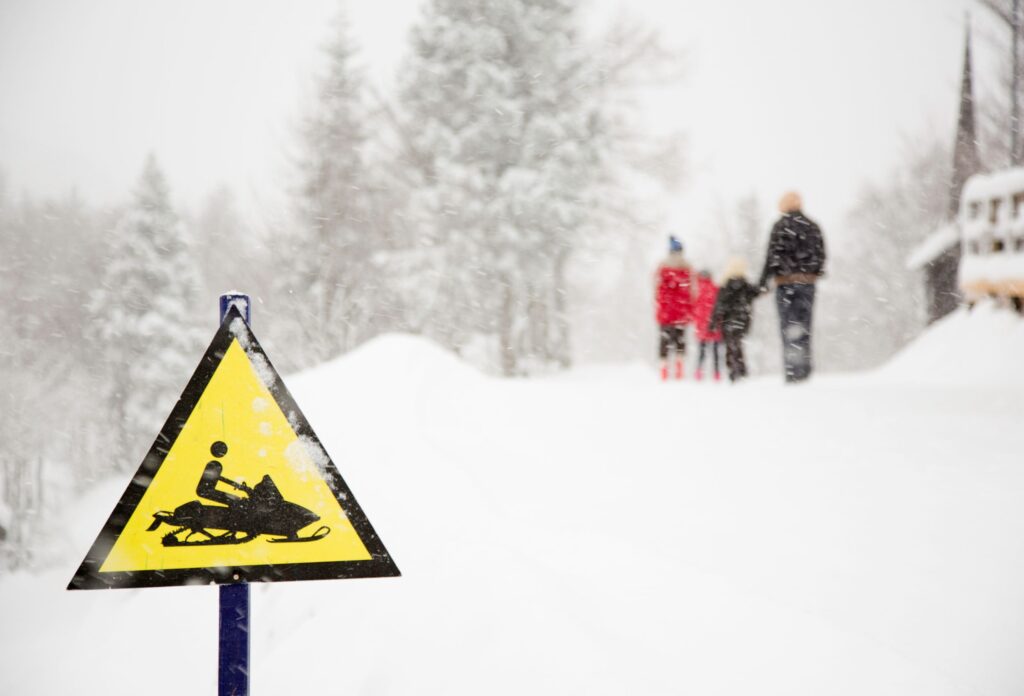 There is a sign posted warning of snowmobiles and in the background it is snowing heavily and a family with children is standing in the background