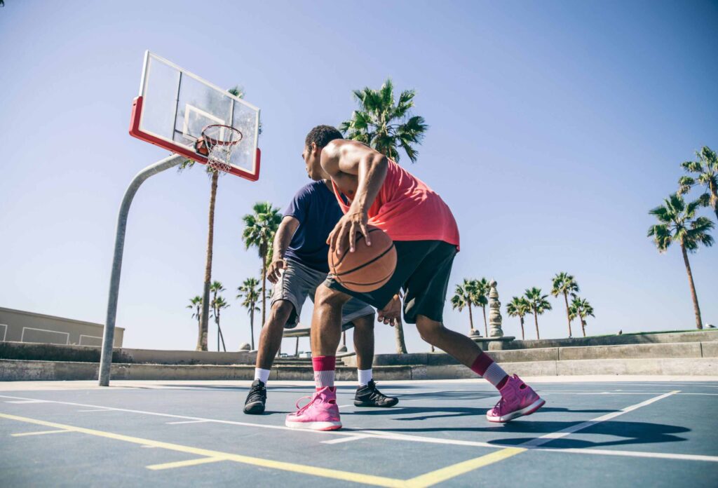 photo of 2 guys in blue and red playing street basketball on a sunny day with palm trees in the background