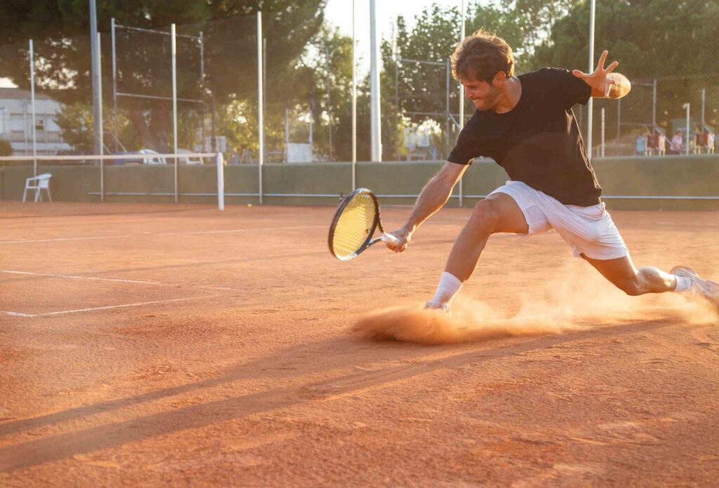 A tennis player in white shorts and black T-shirt makes a sharp lunge to kick the ball so that the ground flies from under his sneaker